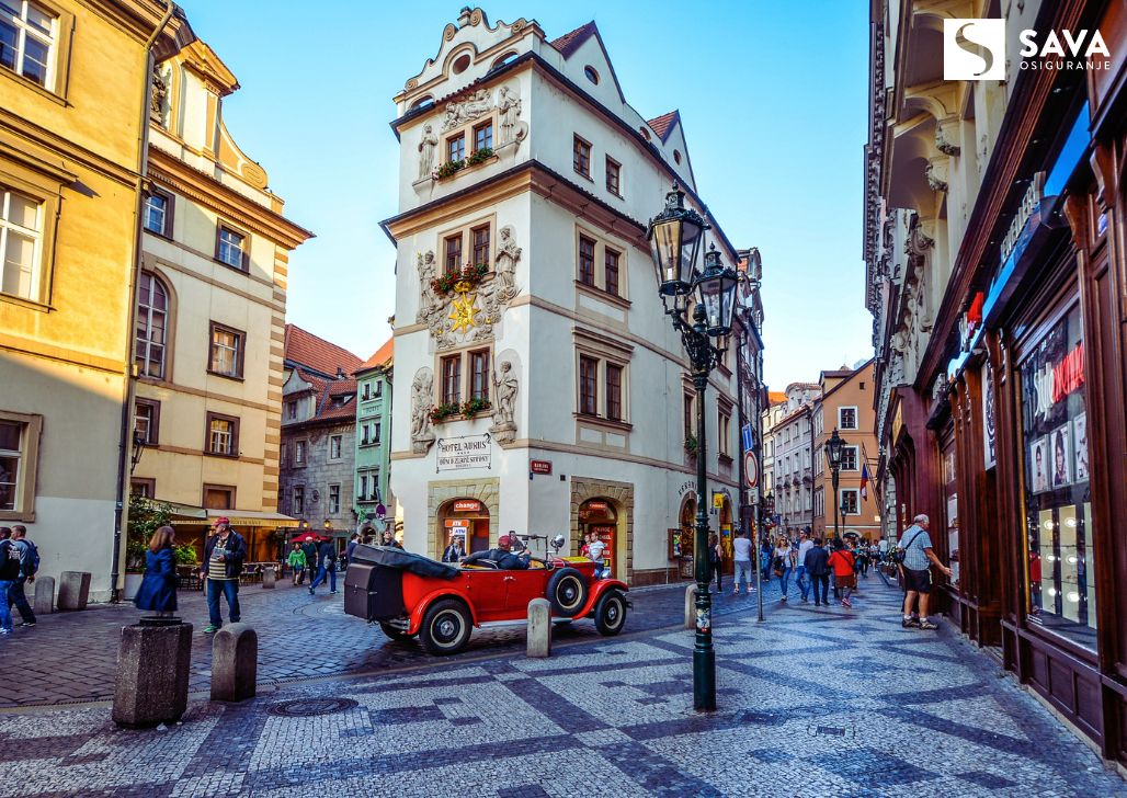 Šarmantna intersekcija u Pragu sa klasičnim automobilom i baroknim fasadama, oživljavajući duh starog grada.