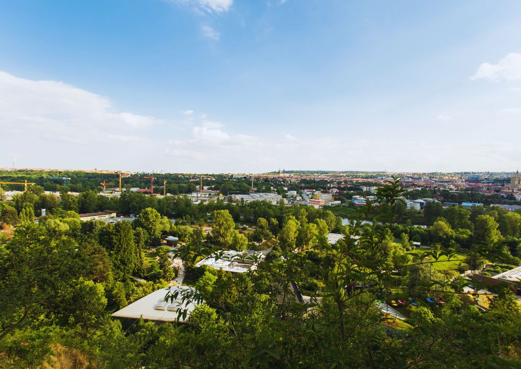 "Prostrani pogled na praški zoološki vrt, smešten u zelenilu grada, sa urbanim pejzažom Praga u pozadini.