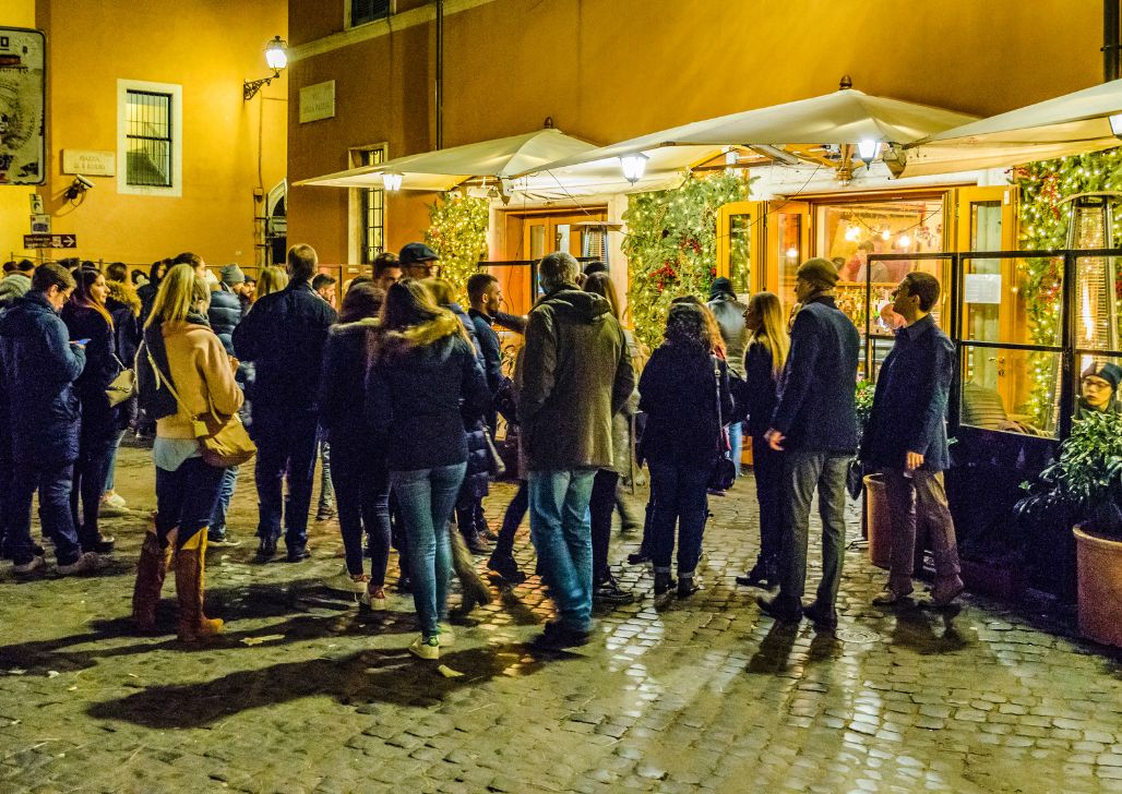 Grupa ljudi okupljena noću u uličnoj atmosferi Rima, okružena svetlima i zgradama