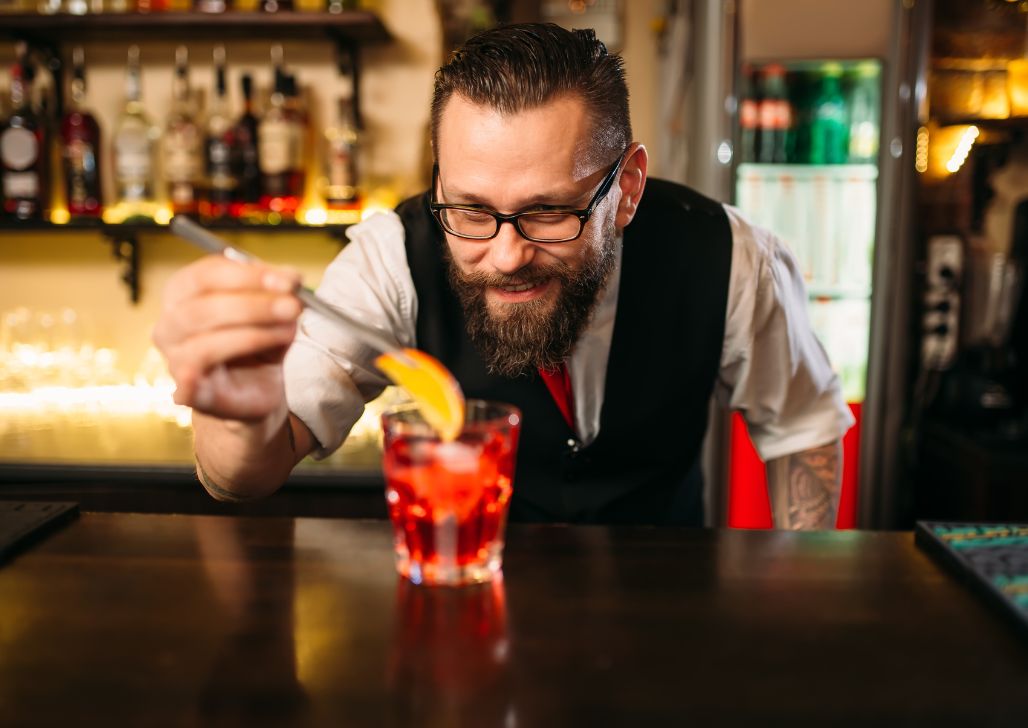 Bartender sa bradom sprema koktel u rimskom baru sa toplo osvetljenim ambijentom.