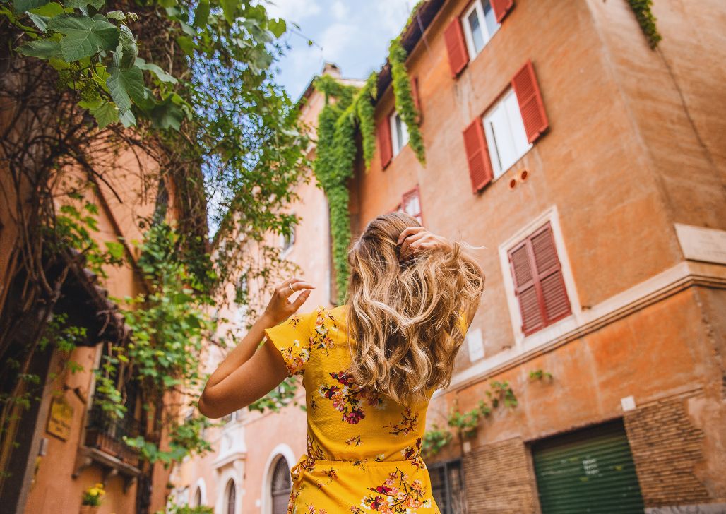 Devojka u žutoj haljini s cvetnim uzorkom šeta popločanom ulicom okruženom istorijskim zgradama u Rimu.