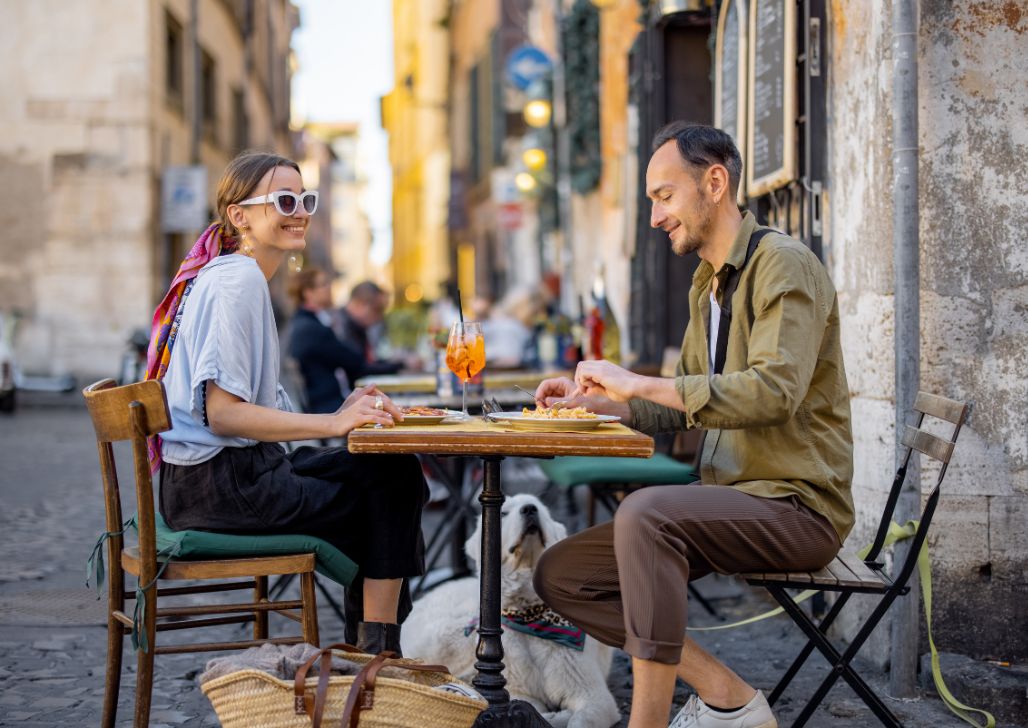 Nasmejani par sedi za drvenim stolom ispijajući aperitive na živahnoj rimskoj ulici, dok ih pas vernik posmatra.