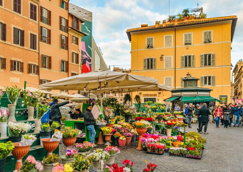 Živopisni cvetni trg sa raznobojnim aranžmanima i ljudima koji šetaju, u istorijskom delu Rima.