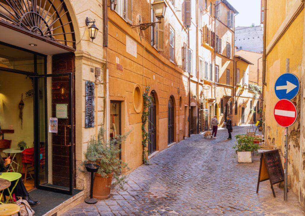 Uska popločana ulica u Rimu, sa istorijskim zgradama i prolaznicima koji uživaju u sunčanom danu.