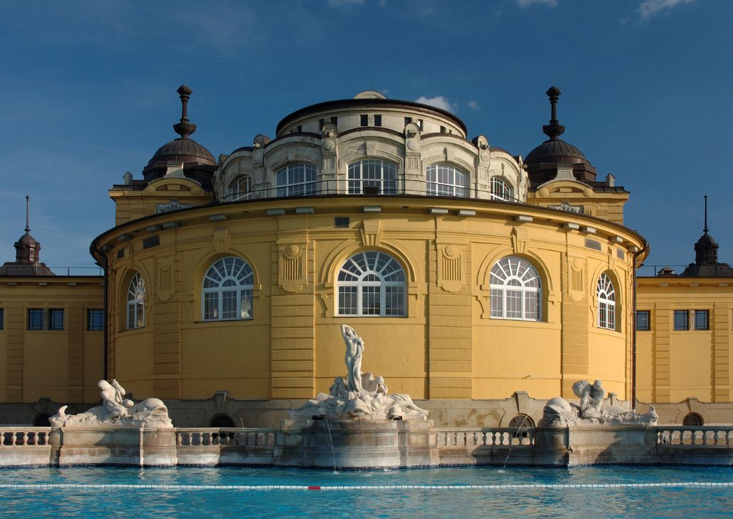 Ikonografska žuta zgrada Széchenyi banje sa skulpturama i plavim bazenom ispred.
