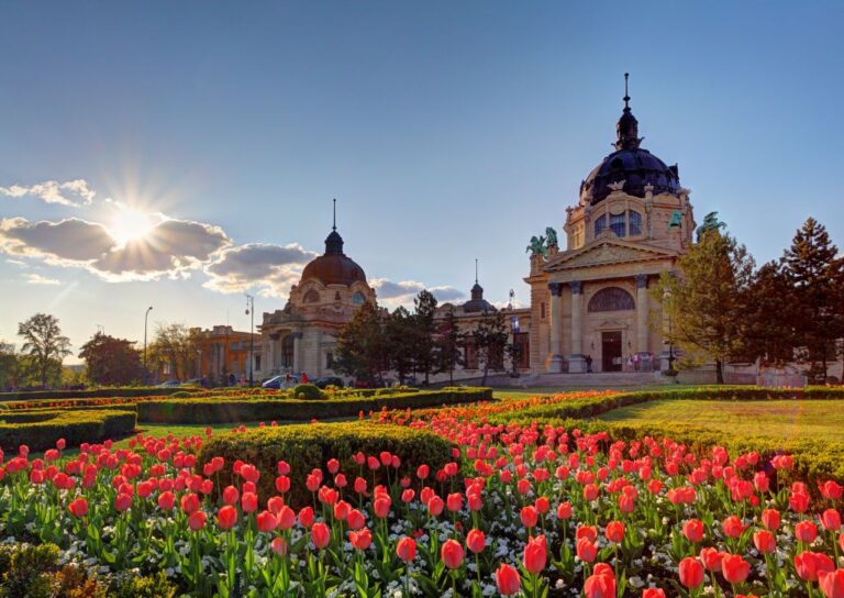 Pejzaž Széchenyi Spa sa cvetnim vrtovima i zgradom spa centra u pozadini pod sunčanim nebom. Najbolje banje u Mađarskoj.